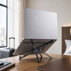 Graphite MagFold adjustable device stand supporting a laptop on a hotel desk for travel work.